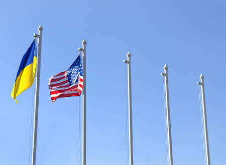 Flagpoles with the flag of Ukraine fluttering next to the flag of USA, against a clear blue skyの写真素材