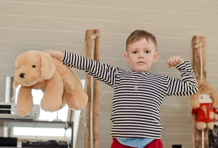 Little boy playfully showing off his muscles as he flexes his arm while holding up a big plush soft...