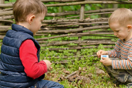 Two young boys kneeling on the ground in the grass in front of an old rustic wooden fence playing with matches as they try to start their own little campfireの写真素材