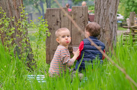 Two little boys sitting drinking at a campsite enjoying a glass of refreshment as the one boy turns to look back at the camera with a mouthful of juiceの写真素材