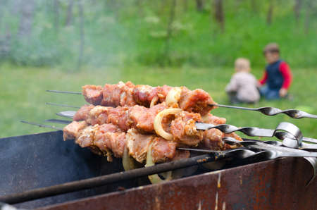 Uncooked meat and onion kebabs piled at the side of s barbecue fire waiting for the coals to be ready for cooking with two children visible in the distance in a lush green campsiteの写真素材