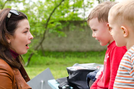 Attractive stylish young woman with her sunglasses on top of her head chatting to two small boys outdoors in a park, close up view of their facesの写真素材