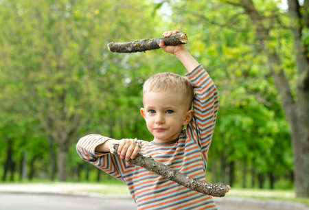 Cute little blond boy wearing camouflage pants standing playing with fighting sticks in a rural road practising his martial artsの写真素材