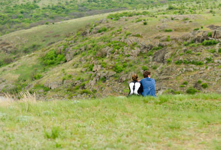 Romantic couple sitting with their backs to the camera on a grassy mountain enjoying the outdoors and the tranquility and solitude of natureの写真素材