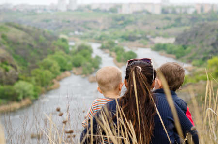 Mother with two small boys enjoying a day in nature sitting in the tall grass on the summit of a hill overlooking a picturesque valley with a wide river with islands, view from behindの写真素材