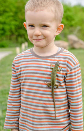 Cute little boy with a live green lizard clinging to his chest on his shirt giving the camera an amused whimsical smileの写真素材