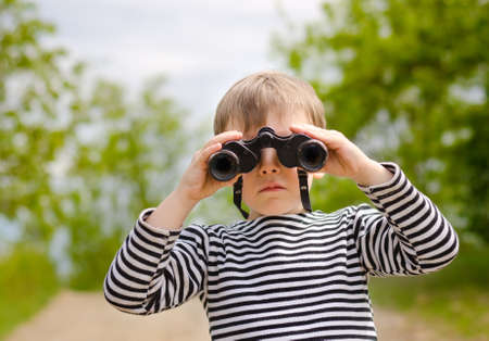 Little boy standing in a green grassy field scanning the surrounding woods with binoculars as he explores the countrysideの写真素材