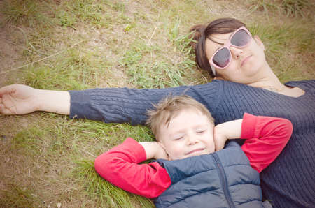 Mother and her young son lying side by side on their backs on the ground with the young boy smiling happily with closed eyes and his mother lying stretched out in her sunglassesの写真素材
