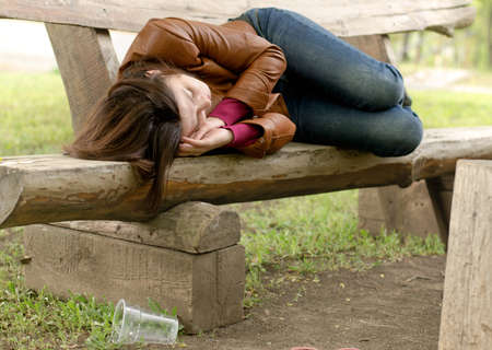 Drunk woman sleeping it off on a wooden bench in a park lying face down in her leather jacket fast asleepの写真素材