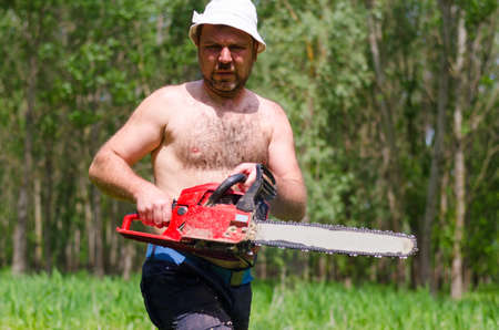 Fit shirtless man wearing a hat carrying a portable petrol chainsaw in woodland as he prepares to fell a tree for fuelの写真素材