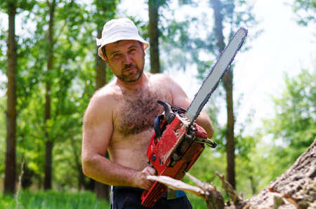 Fit shirtless man wearing a hat carrying a portable petrol chainsaw in woodland as he prepares to fell a tree for fuelの写真素材
