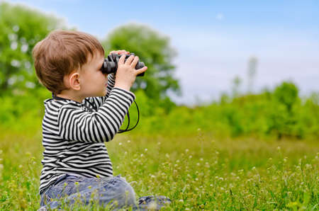 Little boy looking at the camera through binoculars as he kneels in the long grass in a rural meadow, with copyspaceの写真素材