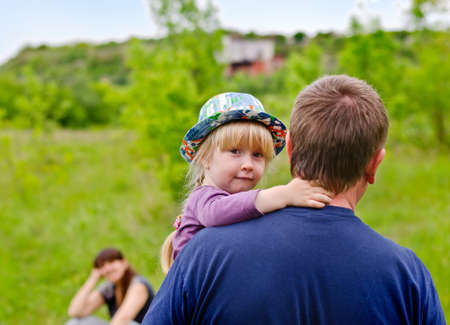 Cute little blond girl wearing a colorful hat being carried by her father looking back over his shoulder at the camera with an uncertain smileの写真素材