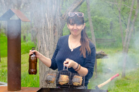 Smiling woman cooking outdoors over a BBQ holding a grill in one hand and a bottle of water to dampen the flames on the hot fire in the otherの写真素材