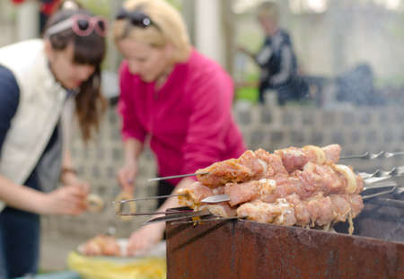 Kebabs ready for cooking standing piled on the grill on an outdoor BBQ as two women continue to prepare more food in the background, focus to the kebabsの写真素材