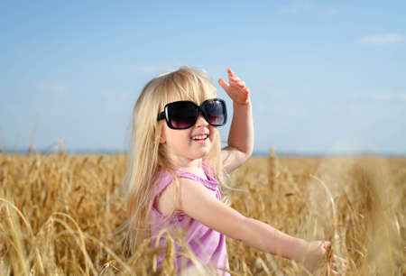 Adorable little blond girl with her hair tied up in pigtails posing in huge fashionable sunglasses in a golden wheat field laughing and wavingの写真素材