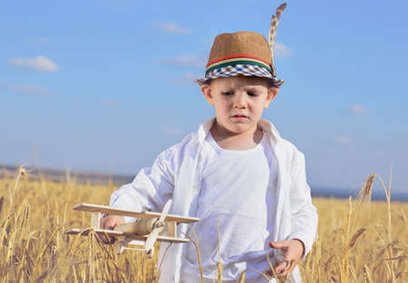 Trendy young boy playing in a field or ripening golden with a wooden toy plane smiling as he imagines himself flying and looping through the airの写真素材