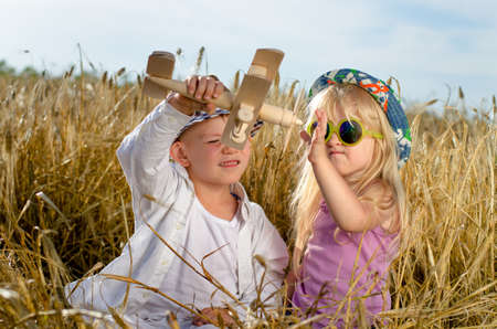 Two young children, a boy and girl in trendy hats, playing together in the summer sunshine with a wooden model plane in a golden wheat fieldの写真素材