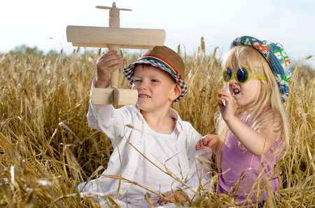 Two young children, a boy and girl in trendy hats, playing together in the summer sunshine with a wooden model plane in a golden wheat fieldの写真素材
