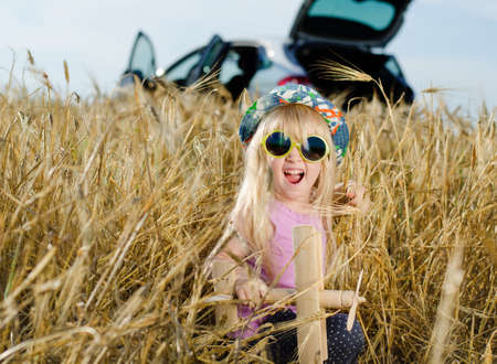 Excited beautiful little blond girl wearing a colorful hat and sunglasses playing with a wooden toy plane in a golden summer wheat field looking at the camera with her mouth openの写真素材