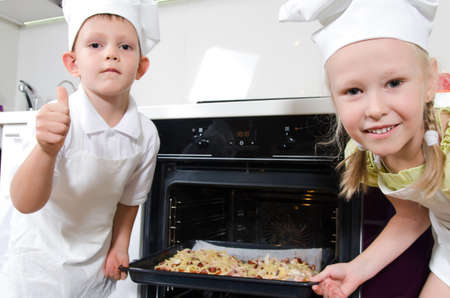 Happy excited young children dressed in white chefs uniforms grinning at the camera as they place a baking tray with homemade pizza in the oven that they have just made themselvesの写真素材