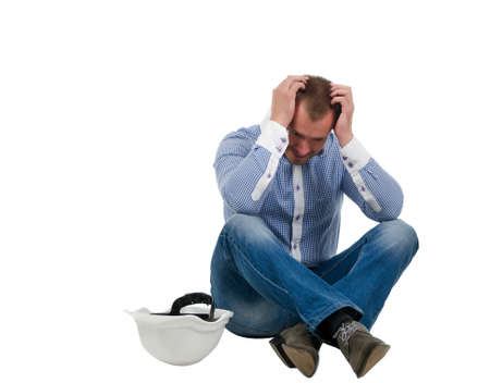 Despairing architect, builder or structural engineer sitting cross legged on the floor with his hardhat alongside him and head in his hands , isolated on whiteの写真素材