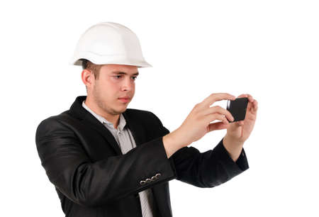 Serious attractive young man in a hardhat and suit taking a photograph using his mobile phone, isolated on whiteの写真素材