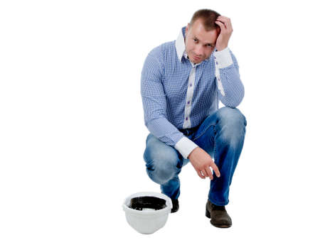 Worried engineer bending down in a crouch with his hardhat in his hand holding his head, isolated on whiteの写真素材