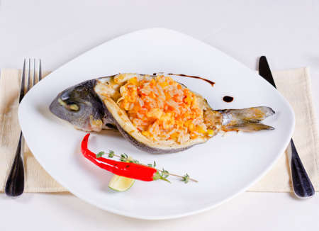Gourmet Sliced Grilled Fish Stuffed with Tasty Risotto Food, Prepared on White Plate with Lemon and Red Chili Pepper. Served on White Table with Fork and Knife on Sides.の写真素材