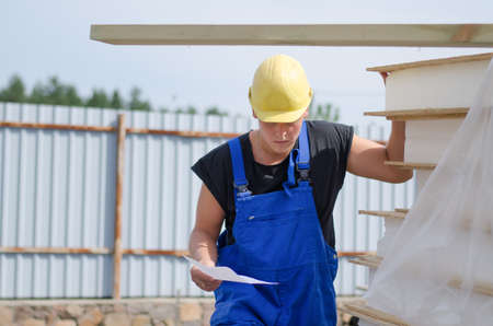 Worried builder in his hardhat and overalls on site with a document in his hand looking at a stack...