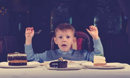 Cute young boy celebrating his birthday with a line up of different cake in front of him pulling a comic face as he tries to make up his mind where to begin eatingの写真素材