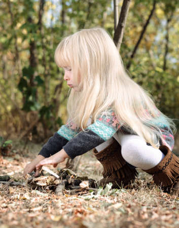 Close up Side View of Little Blond Girl in Autumn Fashion Outfit Playing with Dry Sticks Seriously at the Forest.の写真素材
