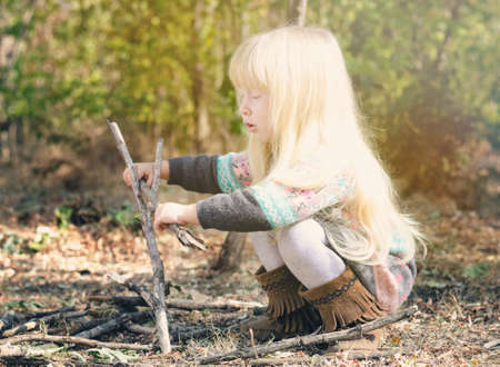 Close up Cute Little Blond Girl Playing with Dry Twigs on Ground at the Woods During Autumn Season. Captured in Side View.の写真素材