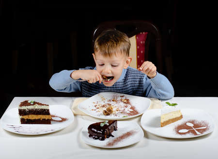 Cute little boy enjoying a treat of party cakes hungrily gulping down a big mouthful with slices of a variety of different cakes displayed in front of himの写真素材