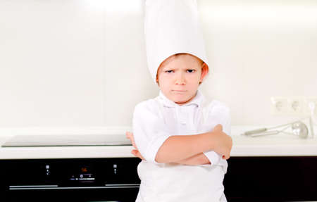 Cross determined little boy chef standing in his white apron and toque in the kitchen with his arms...