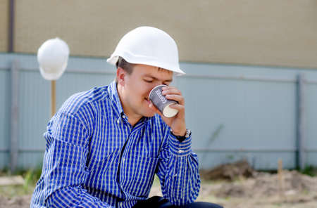 Close up side view of a young workman in a hardhat sipping coffee on a building site while the team...