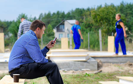Foreman or engineer reading a text message on his mobile phone as he pauses in his work for a coffee break as workmen continue work in the backgroundの写真素材