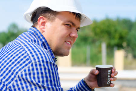 Close up side view of the face of a young builder in a hardhat sitting on a building site taking a coffee breakの写真素材