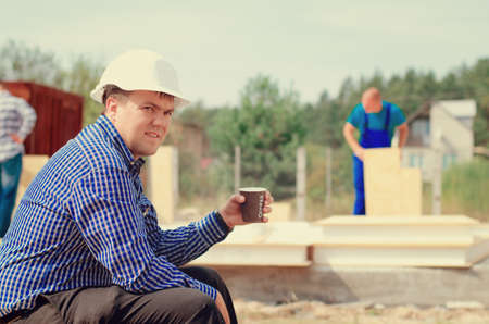 Site foreman taking a coffee break as the workmen continue working behind him erecting wall panels,...