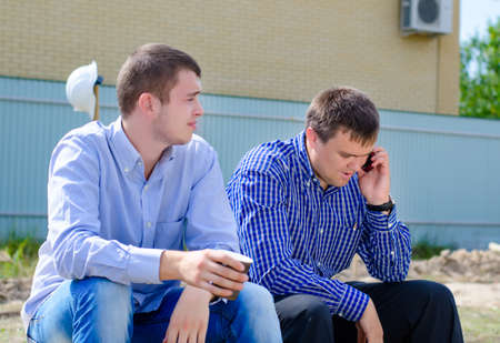 Two architect partners taking a coffee break on a building site with one sipping coffee while the other chats on his mobileの写真素材