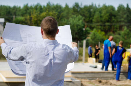 View from behind of a young architect or engineer checking a building plan on site holding it open in his hands as workmen work in the backgroundの写真素材