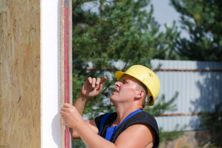 Builder ensuring that a beam is vertical holding a builders level against the wooden panel as he...