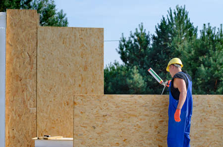 Builder applying glue to an insulated wooden wall panel on a building site for a new build house