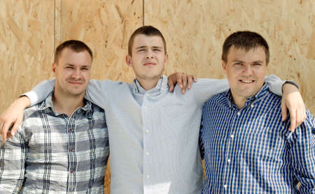 Three male friends standing arm in arm in front of a wooden wall panel smiling happily at the camera as they enjoy the camaraderie of being togetherの写真素材