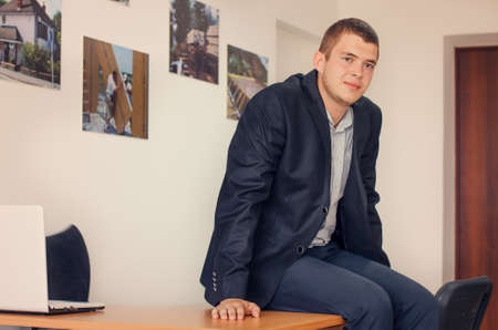 Portrait of Young Businessman Wearing Business Casual Attire Sitting on Desk in Office and Looking Optimisticの写真素材
