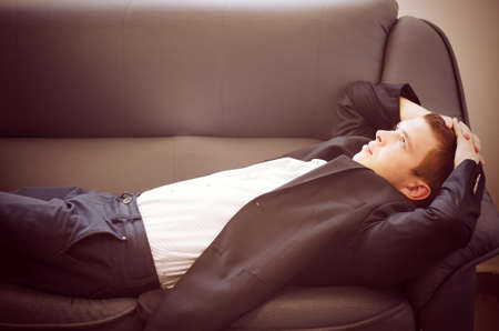 Young Man Lying on Sofa Relaxing with Hands Behind Head Looking Off Into Space as if Daydreamingの写真素材