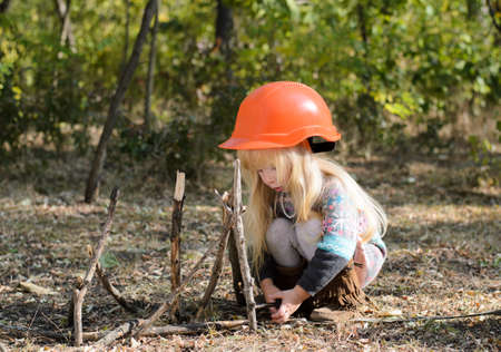 Close up Serious Little Blond Girl with Orange Helmet Playing with Sticks on a Grassy Ground at the Woodland.の写真素材
