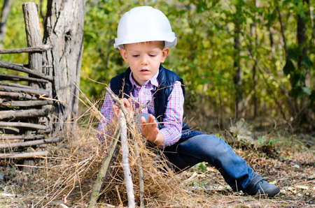 Close up Sitting Little Young Boy in Autumn Clothing with White Helmet Playing Sticks on a Grassy Groundの写真素材