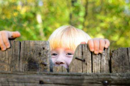 Close up Playful Little Blond Girl Peeking on Old Wooden Fence at the Gardenの写真素材