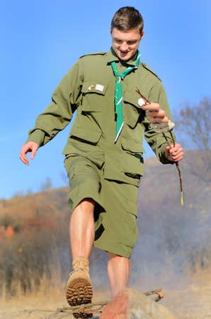 Boy Scout Wearing Uniform Holding Stick of Uncooked Sausages and Breaking Wood for Campfire